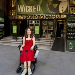 Wheelchair user in a red dress outside the Apollo Victoria Theatre, with Wicked signage and an accessible ramp.