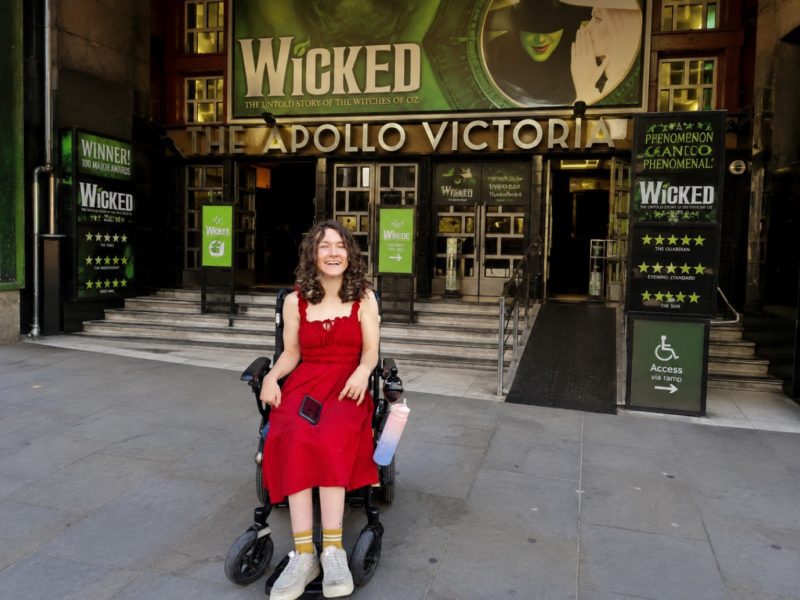 Wheelchair user in a red dress outside the Apollo Victoria Theatre, with Wicked signage and an accessible ramp.