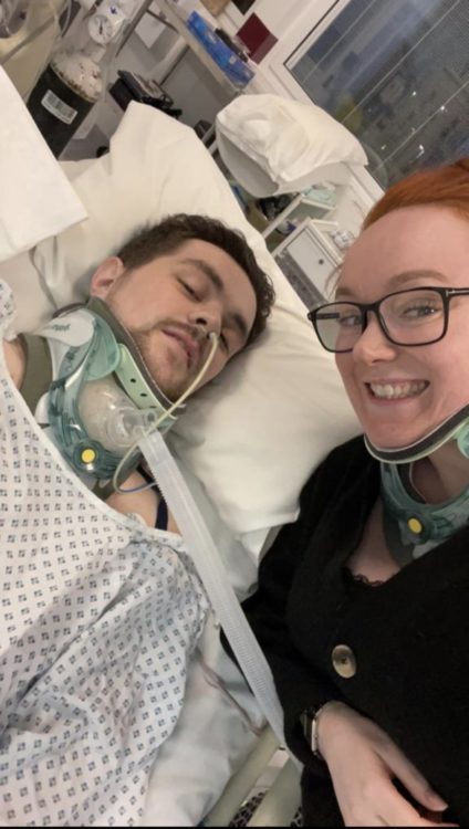 Johkes rests in a hospital bed with breathing support as a smiling woman takes a selfie beside him.