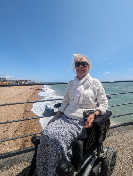 Clare, a power wheelchair user and inventor of Trabasack, on a seafront promenade with the beach and sea behind her.