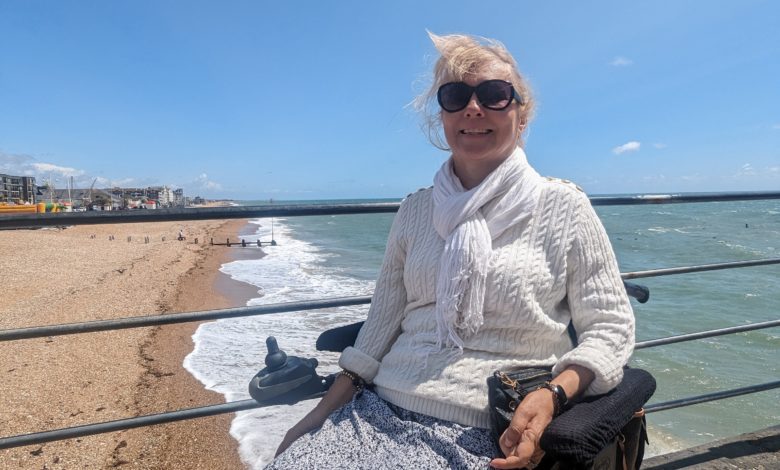 Clare, a power wheelchair user and inventor of Trabasack, on a seafront promenade with the beach and sea behind her.