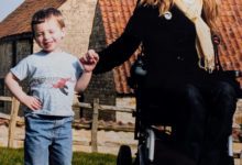 A smiling woman in a wheelchair holds hands with a young boy on a sunny lawn in front of rustic farm buildings.