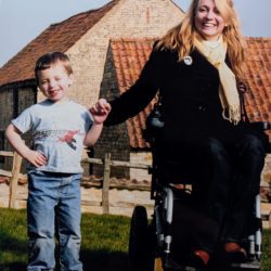 A smiling woman in a wheelchair holds hands with a young boy on a sunny lawn in front of rustic farm buildings.