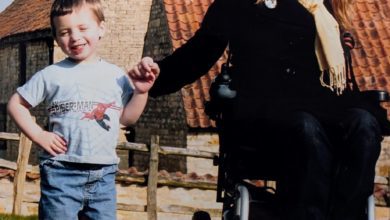 A smiling woman in a wheelchair holds hands with a young boy on a sunny lawn in front of rustic farm buildings.