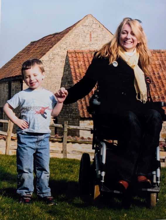 A smiling woman in a wheelchair holds hands with a young boy on a sunny lawn in front of rustic farm buildings.