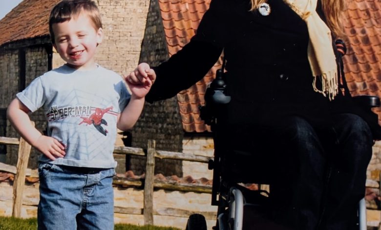 A smiling woman in a wheelchair holds hands with a young boy on a sunny lawn in front of rustic farm buildings.