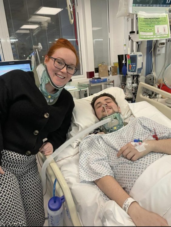 Johkes smiles from a hospital bed beside a smiling woman, both wearing neck braces amid medical equipment.