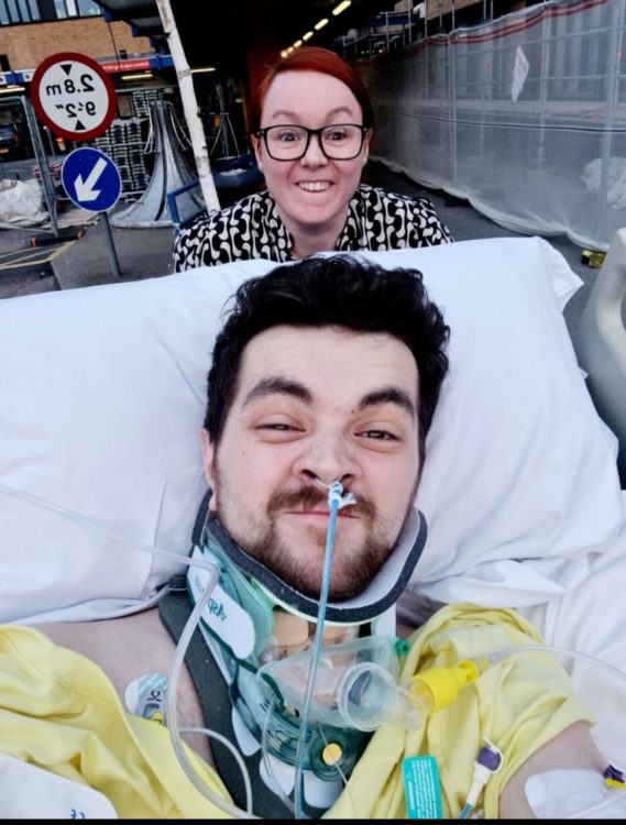Johkes lies in a hospital bed with breathing support while a smiling woman stands behind him outdoors.