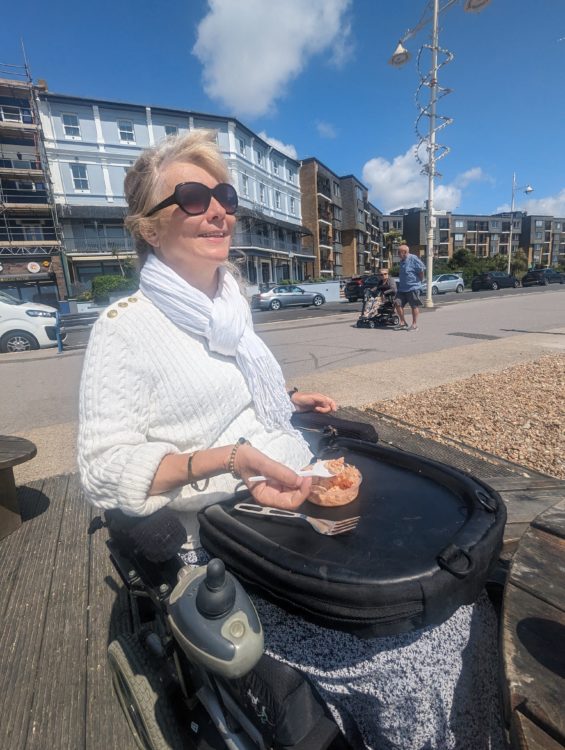 Clare, a power wheelchair user, eating on the seafront with a Trabasack lap tray across her wheelchair.