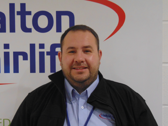Man wearing a Halton Stairlifts uniform stands smiling in front of a large Halton Stairlifts company sign.