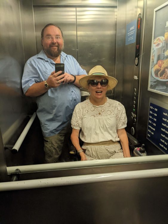 Duncan and Clare Edwards smiling in a mirrored passenger lift. Clare is seated in her wheelchair, wearing a sunhat and sunglasses, while Duncan takes the photograph.