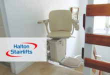 Cream-coloured stairlift with a fold-down footrest installed at the top of a staircase, beside a wooden banister.
