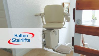 Cream-coloured stairlift with a fold-down footrest installed at the top of a staircase, beside a wooden banister.
